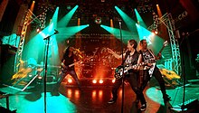 The band Speed limit playing on a brightly teal lit stage with the bass-player on the left, two guitarists on the right in the foreground and drums in the back.