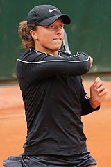 A woman wearing a black cap and black jacket has swung her right arm, which is holding a tennis racquet, around her left shoulder, as she looks into the distance in front of her.