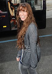 A picture of a woman who is smiling. She is walking on a street and she is looking back over her left shoulder. She wears a grey sweater with some black frames, and jeans of the same colour. In the background a black bus is visible.