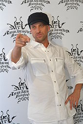 A white man in a white shirt with sleeves rolled up and a black baseball cap. He is pointing at something in front of him with his right hand.