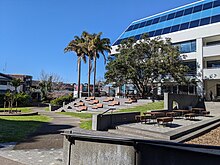 A picture of a park, with grass and path moving through. On the right are many picnic benches and tiered steps with lounge chairs.