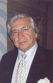 A man with gray hair is wearing glasses and a black tuxedo