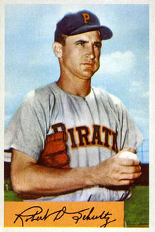 A man in a gray baseball jersey with "Pirates" written on the chest and a navy blue cap with an orange "P" on the front stands with a glove under his arm and a ball in his hand.