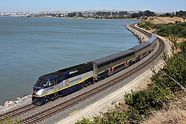 A passenger train along a shoreline. The diesel locomotive is gray with a navy blue underside, yellow bottom stripe, black roof, and navy blue cab area. The bilevel passenger cars are stainless steel with a black upper window stripe and a navy blue lower window stripe with a yellow pinstripe.