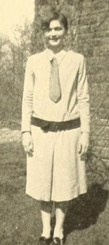 A smiling young white woman, standing outdoors, wearing a light-colored dress with long sleeves and a necktie