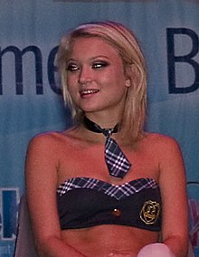 A blonde white woman in a bikini top and decorative necktie is looking off to the camera's left and slightly smiling.