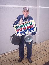 Full body shot of fan Freddy Sez, holding a pan with a shamrock and a sign that says "SCREAM-WHISTLE, KEEP UP NOISE!, IT HELPS YANKS!".