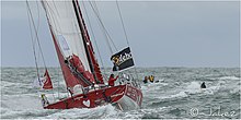 Voilier rouge et blanc, vu de l'arrière, sur l'océan. Le skipper se tient à l'arrière.