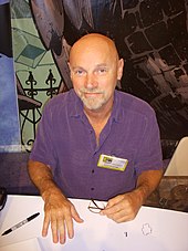 Jim Starlin, a bald man with a goatee wearing a purple shirt, sits at a table.