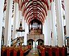 Interior of the church, view to the organ, white walls and columns, red accents in the vault, wooden benches, pulpit on the left