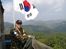 A uniformed soldier leans against the railing of a guard post overlooking the mountains. There is a Korean flag behind him.