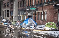 A snowy street in the foreground, a line of tents on ppthe sidewalk and a brick building in the background.