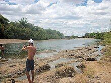 Blick auf den Flusslauf mit zwei Männer in Shorts vorne seitlich im Bild mit nacktem Oberkörper, dahinter begrünter und teilweise versandeten Flussufer rechtsseitig und bewölktem aber blauem Himmel am Horizont in Farbe.