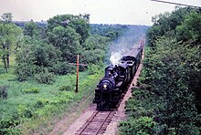 A small black steam locomotive pulling a passenger train down a single-track mainline, with trees in the background