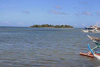 Small low green island in blue sea, at about 600 m, with several palm trees on top. To right prows of four bangkas – local boats for fishing or diving.