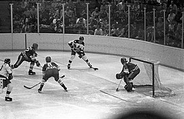 Black and white in-game action photo including a goaltender, two defenders, and two attacking forwards