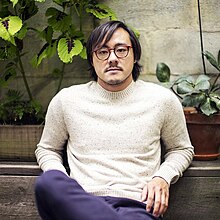 Portrait of the writer Brian Lam taken in 2014. Mr Lam is an ethnically Chinese man who is wearing a white long-sleeve shirt and dark blue pants. In this portrait, he is seated, reclining slightly in front of a low wall, on which he is resting both elbows. He is wearing glasses and a thin beard and moustache, and his hair, cut to approximately eyebrow length, is parted on the left. In the background are two potted plants.