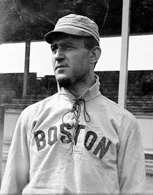 A man wearing a baseball cap and jersey with "Boston" written across the chest shown from the waist up looks to the left