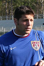 The head and torso of a man in his 30s, wearing a blue jersey. On his chest is a crest, featuring the letters "US" and a picture of a football (soccer ball).
