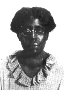 An African-American woman with short hair, wearing glasses and a dress with a ruffled collar