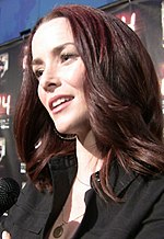 A 31-year-old woman with long, brown hair, smiling with her head tilted at someone holding a microphone to the left of the camera.