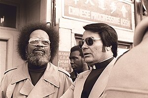 Cecil White and Jim Jones, both wearing trench coats, stand together in front of a doorway.