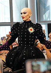 Sasha Velour trägt eine Glatze, lila Lidschatten, roten Lippenstift, falsche Wimpern, Wimperntusche, aufgemalte Augenbrauen, ein schwarzes Kleid mit silbernen Kugeln, auf der linken Brust eine gelbe, blumenförmige Brosche mit rotem Kern, lila Handschuhe sowie am linken Ohr eine Kette, an der eine goldene Blüte und ein Bernstein hängen. Im Hintergrund sind Fenster und mehrere Personen zu sehen, von denen einige Smartphones hochhalten.