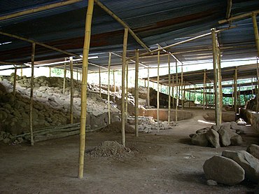 A cleared area of dirt strewn with rubble, with raised areas to the left and right and covered by a roof supported by wooden poles.