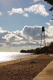 Photo of a sandy beach, a lake on the left, a boardwalk on the right, and trees and a water tower in the distance