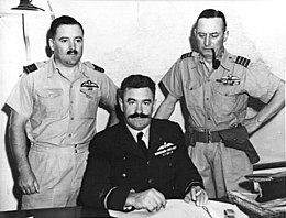 Half length portrait of three military men behind a desk, all with pilot's wings on left breast pocket. One of the men, seated, has a large dark moustache and is wearing a dark winter uniform. The other two, standing on either side of the seated figure, wear short-sleeved tropical uniforms; one of them has a small moustache, the other has a holster on his belt and is clean-shaven and smoking a pipe