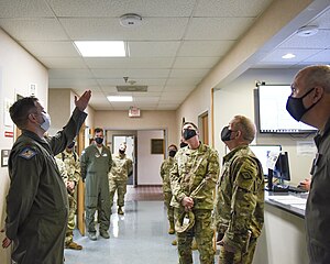 National Guard soldiers standing in circle gesture at small white device mounted to ceiling in office-like military facility.