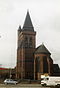 A church tower topped with a pyramidal spire and a round stair turret behind; to the right is an apse with buttresses; a car and a white van pass in front