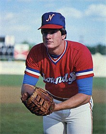 A man wearing a red baseball jersey with "Sounds" written in white and blue across the chest and a blue cap with an "N" on the front in white and red leans forward, hands in baseball glove, as if looking to his catcher for the sign.