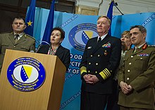 A woman in a dark suit speaks behind a wooden lectern, while three men in military uniforms, and one woman, stand to her sides.