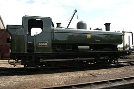 A pannier tank locomotive is standing outside an engine shed. The locomotive is mainly painted green above the running plate, although the chimney is black. On the side of the pannier tank a yellow circle encloses the yellow letters "G W R". The G and are shaped to match the curve of the enclosing circle.