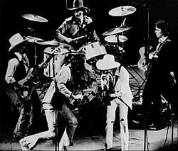 A group of men performing on a stage. One is playing a violin, three are playing guitars, and one is playing drums. The drummer and two of the guitarists are wearing cowboy hats.