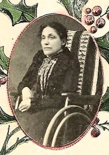 A white woman with dark hair, wearing black, seated in a wheelchair with a high back. Her hands are clasped in her lap. The photograph is in an oval frame decorated with holly leaves and berries.