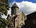 Petit monastère en pierres roses et grises incluent par une tour surmontée d'une croix, sur la gauche un bâtiment est étançonné par des échafaudages en bois.