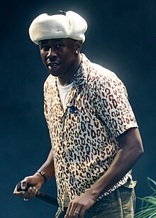 A man in a soft white mushroom hat and panther-print dress shirt is blinged out as he holds the mic at waist-level and looks at the camera.