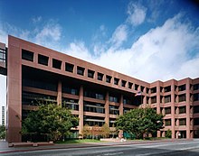 A rectilinear four-story brown finished stone building with many recessed square windows and a large recessed entry area. An American flag flies from a pole in front