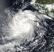 A photograph of a powerful hurricane off the Pacific coast of Mexico. It has a small and clear eye surrounded by a thick ring of solid white clouds, indicating intense convection. There are several well-pronounced rainbands, especially on the left side of the hurricane. Also on the left side of, and perpendicular to, the hurricane are some thin streaks of high clouds jutting out westward; these are indicative that the hurricane has healthy outflow, which is one of the things a tropical cyclone needs to get stronger and stay strong.