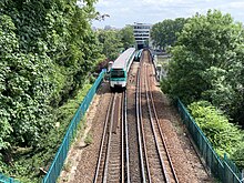 Photographie d'une rame de métro vue de face sur une voie ferrée aérienne bordée d'arbres buissonnants.