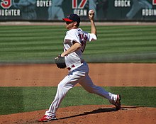 A man in a white baseball uniform and navy-blue baseball cap with a red brim throws a baseball with his right hand from atop a dirt mound on a grass field