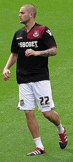 A young man wearing a black and claret shirt, white shorts and white socks, standing on a grass field