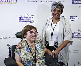 A photograph of Judy Heumann in her power chair next to Barbara Ransom. They are holding hands and smiling, standing in front of a sponsor banner.