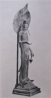 Three-quarter view of a very slim and tall statue carrying a vase in with two fingers of her left hand. Her right arm is bend with the palm of her right hand facing upward. A halo on a pole is seen behind the statue. Black and white picture.