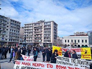 Κόσμος συγκεντρωμένος σε πλατεία, με πανό. Το μπροστινό γράφει «Όχι άλλοι νεκρό στο βωμό του κέρ[δους]. Οι ιδιωτικοποιήσεις...».»