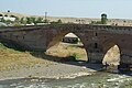 Pont en briques de teinte rosâtre dont on voit deux arches en arc brisé, la première couvre une rivière, la seconde un chemin en pierres partiellement recouvertes par de l'ivraie ; quelques arbres sont visibles au-delà du tablier du pont.