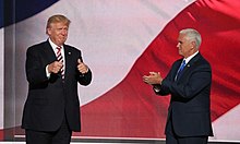 Donald Trump and his running mate for vice president, Mike Pence. They appear to be standing in front of a huge screen with the colors of the American flag displayed on it. Trump is at the left, facing toward the viewer and making "thumbs-up" gestures. Pence is at right, facing Trump and clapping.
