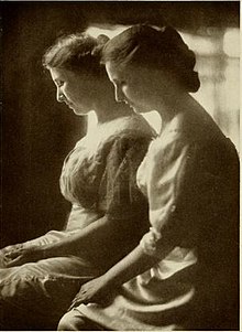 Black and white image of two women with their heads bowed and hands in their lap. They're both wearing dresses and have their hair pinned back.